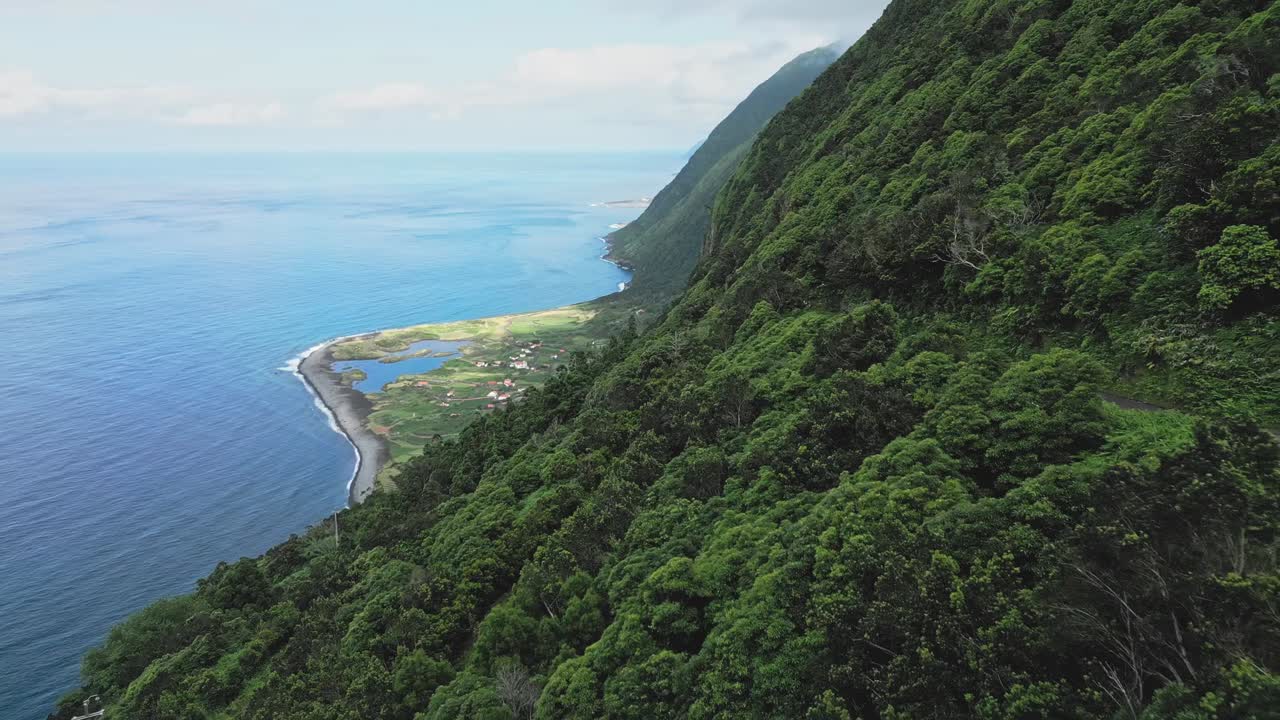 Flycam kéo lùi chậm từ vách đá xanh tươi phía trên Fajã da Cumbre, đảo São Jorge, Azores, Bồ Đào Nha, ngôi làng ven biển, đầm phá và Đại Tây Dương xanh biếc.