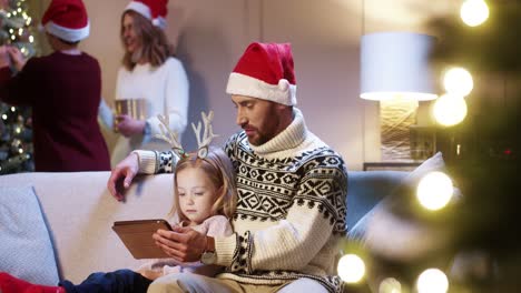 Portrait-Of-Caring-Dad-In-Santa-Hat-With-Cute-Little-Girl-Sitting-In-Room-Tapping-On-Tablet-Buying-Xmas-Gifts-On-Internet-Mom-And-Son-Decorating-Glowing-Christmas-Tree-Family-Holiday-Concept