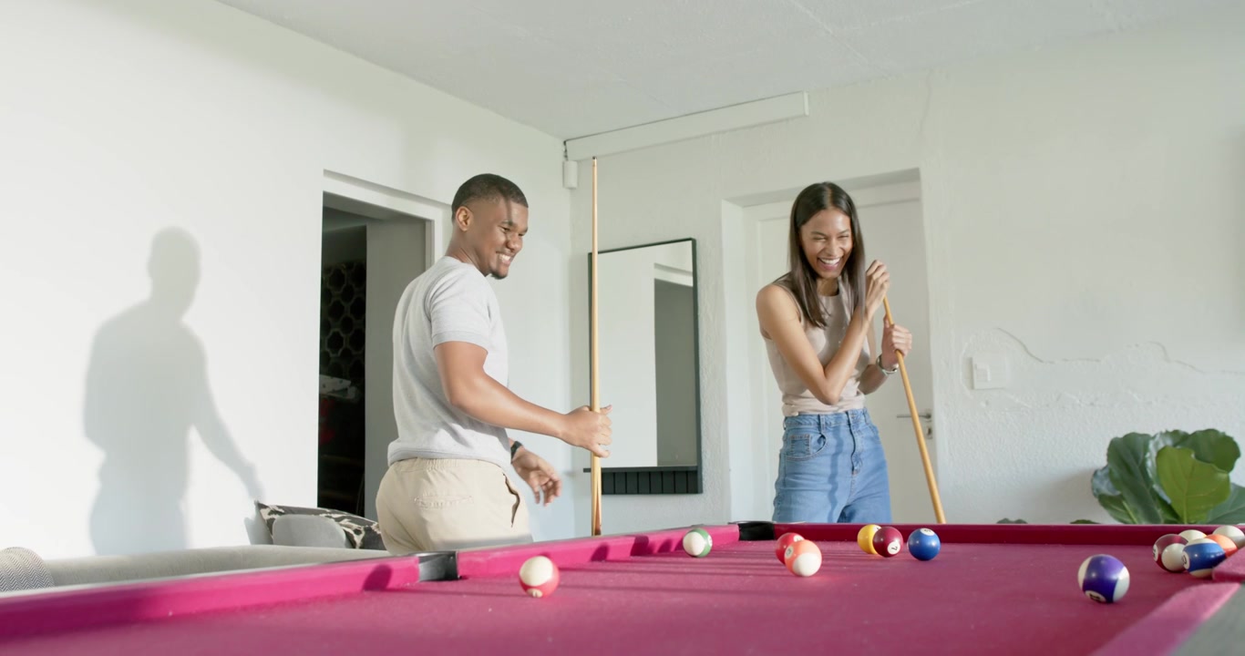 African American couple laughing, man tapping cue at magenta pool table preparing to play at home