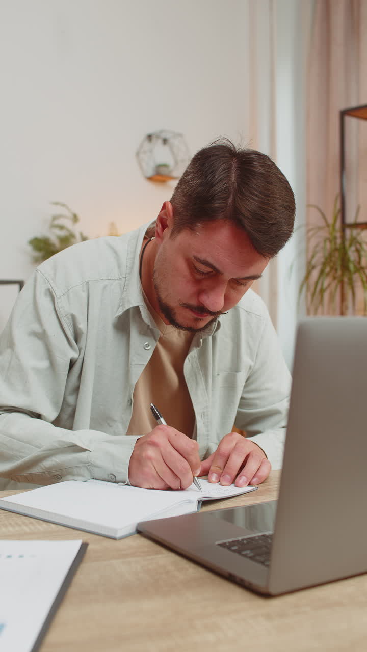 Young man writing down notes while attending online office meeting on laptop at home office desk