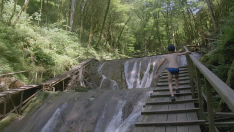 boy walking on wooden walkway over a waterfall in forest