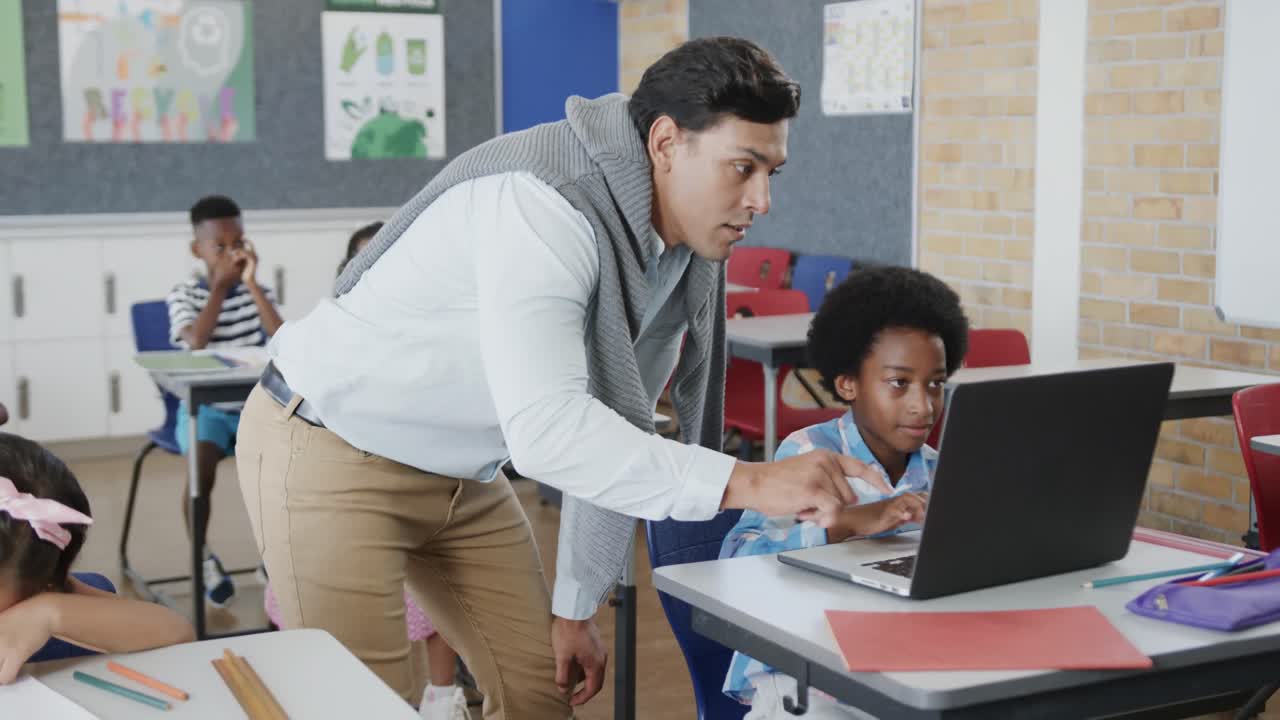 un maestro feliz y diverso ayudando a un niño en el escritorio usando una computadora portátil en la clase de escuela primaria, cámara lenta