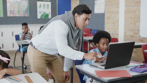 Happy-diverse-male-teacher-helping-boy-at-desk-using-laptop-in-elementary-school-class,-slow-motion