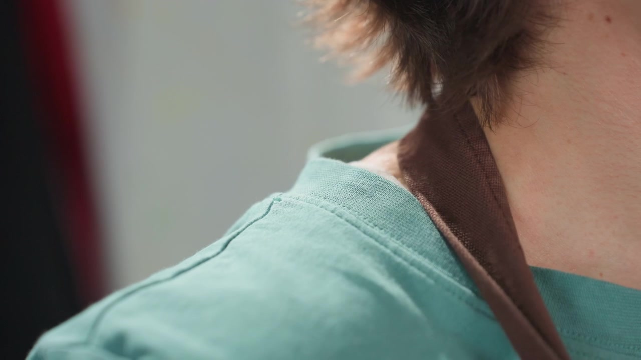 Primo piano della spalla di un vasaio bianco in uno studio di ceramica, luce soffusa sul collo, camicia color ottanio e spallina del grembiule visibili, pausa contemplativa, dettaglio intimo dell'artista che si prepara per una sessione di ceramica.