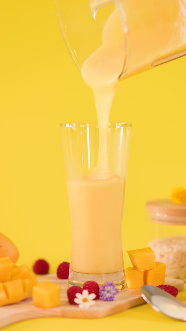 Un frullato denso di mango si versa lentamente in un bicchiere alto con guarnizione di frutta tropicale, illuminazione brillante da studio.
