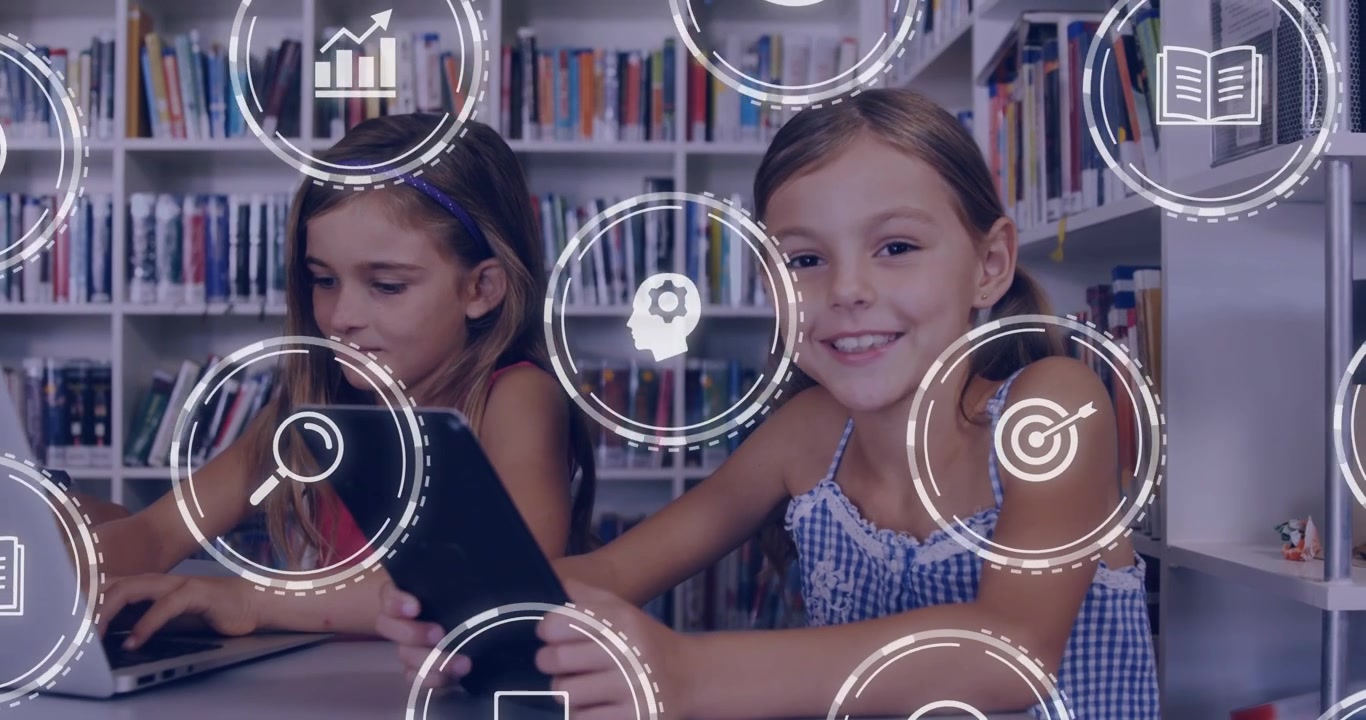 Two female child friends sitting at table in library using tablet and laptop showing learning icons
