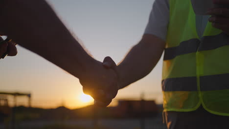 portrait of hands of two builders. builder shaking hand the builder on built house background. close up of a handshake of two men in green signal vests against the background of the sun