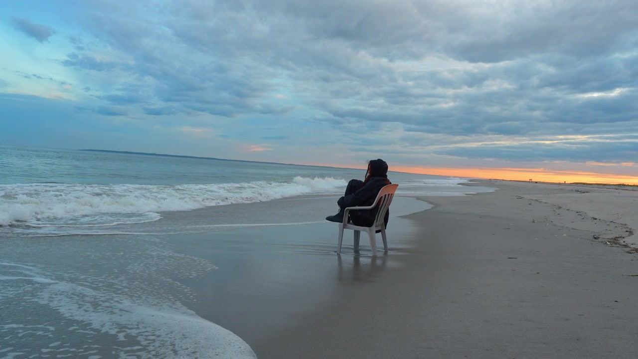 Person sitting in a chair at the ocean water line at sunset. Foam washes around the base of the plastic seat as waves collapse along the broad sandy shore.