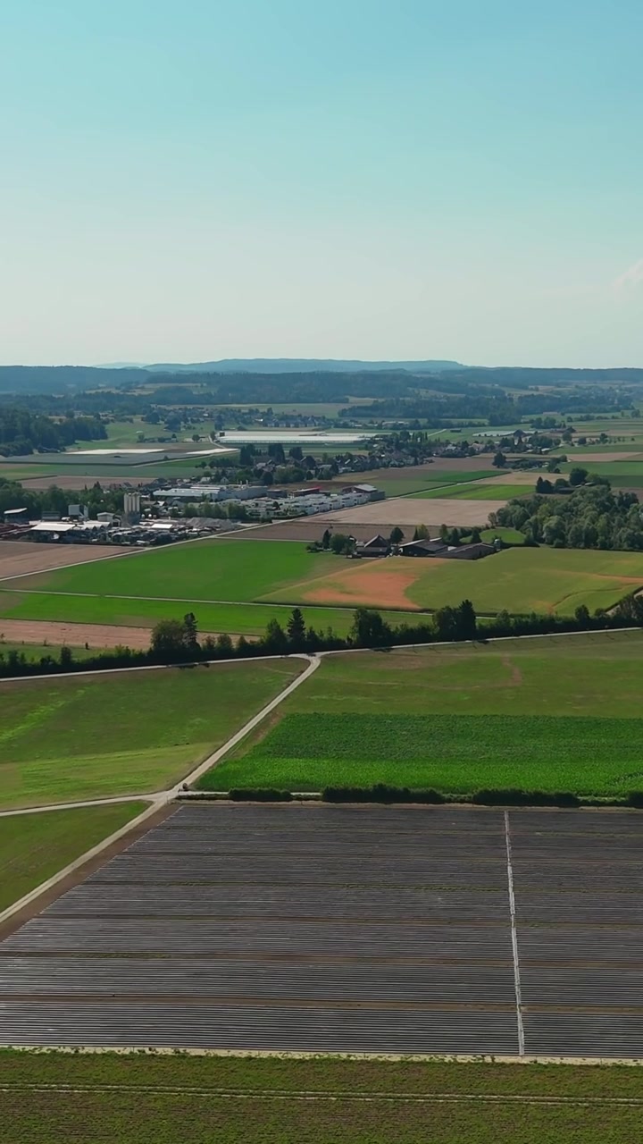 Vertical aerial panorama over Swiss farmland, showing geometric crop fields, country roads, scattered buildings and rolling countryside stretching toward a distant horizon.