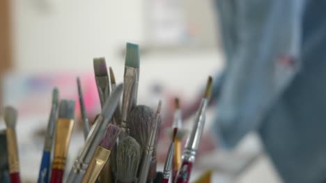 pots of artists brushes in shallow depth of field, behind an artist works in home studio out of focus