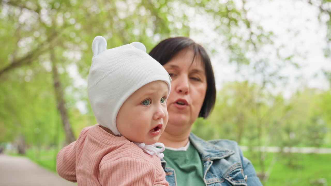 Woman Strolling With Baby Nearby Trees, Mother And Infant Enjoying Peaceful Walk Amidst Trees In Park, Woman Carrying Her Baby Along Path Surrounded By Trees During Tranquil Spring Day