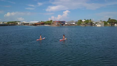 aerial over two women rowing paddleboards towards a coastal village in honduras