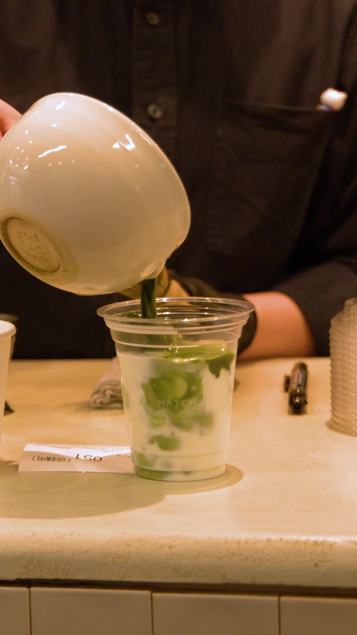 A barista pours concentrated green matcha tea from a ceramic bowl into iced milk.