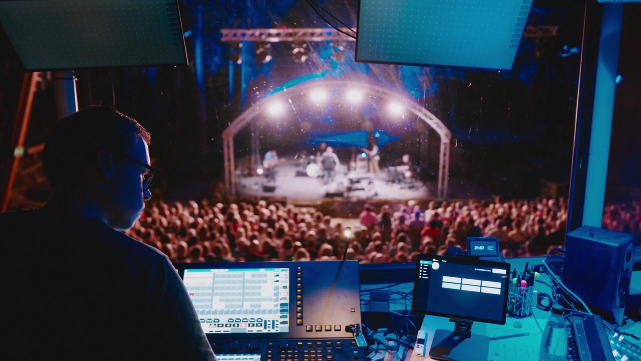 Handheld, Sound engineer works at a digital mixing console while monitoring musicians performing live on stage, showing behind the scenes audio production at a concert