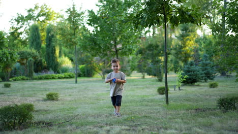 little kid wearing grey t-shirt running by the grass following his ball. toddler boy picks a toy and approaches the camera happily.