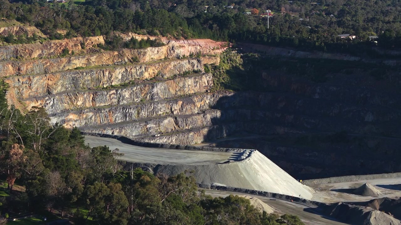 Cinematic aerial footage of Montrose Quarry in Melbourne, revealing steep terraced rock layers, machinery and mounds under clear daylight. Vivid, detailed landscape in industrial setting.