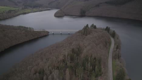 drone shot in luxemburg near the river lac de la haute-sure at a nature park öewersauer on a cloudy day with cars driving on the road near the forest and trees and a bridge in the background log