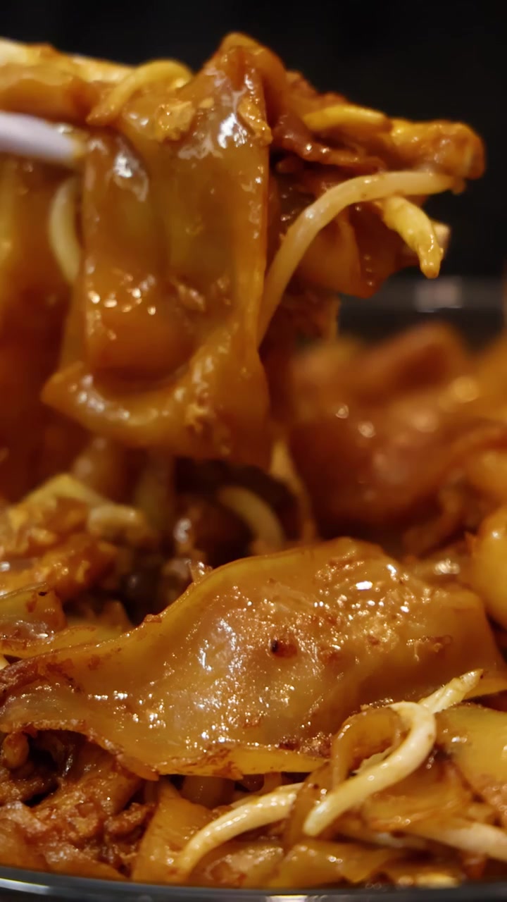 Primo piano di bacchette che raccolgono noodles di riso piatti saltati in padella con salsa, uovo e germogli di soia sotto un'illuminazione calda, evidenziando la consistenza lucida e l'attrattiva del cibo da strada.