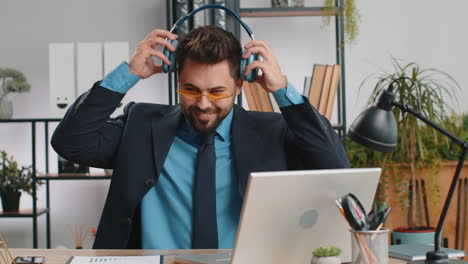 hombre de negocios caucásico con auriculares escuchando música bailando después de trabajar en la computadora portátil en la oficina