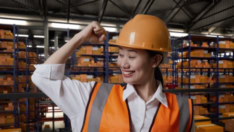 primer plano de una ingeniera asiática con casco de seguridad de pie en el almacén con estantes llenos de productos de entrega. flexionando su bíceps y sonriendo a la cámara en el almacenamiento