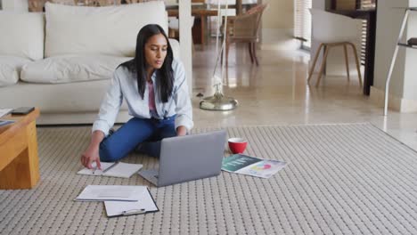 mujer de raza mixta usando computadora portátil pasando por el papeleo bebiendo café trabajando desde casa