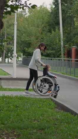 mother carefully lowers wheelchair with daughter on road on spring day. woman and girl with chronic health condition head home after walking in park