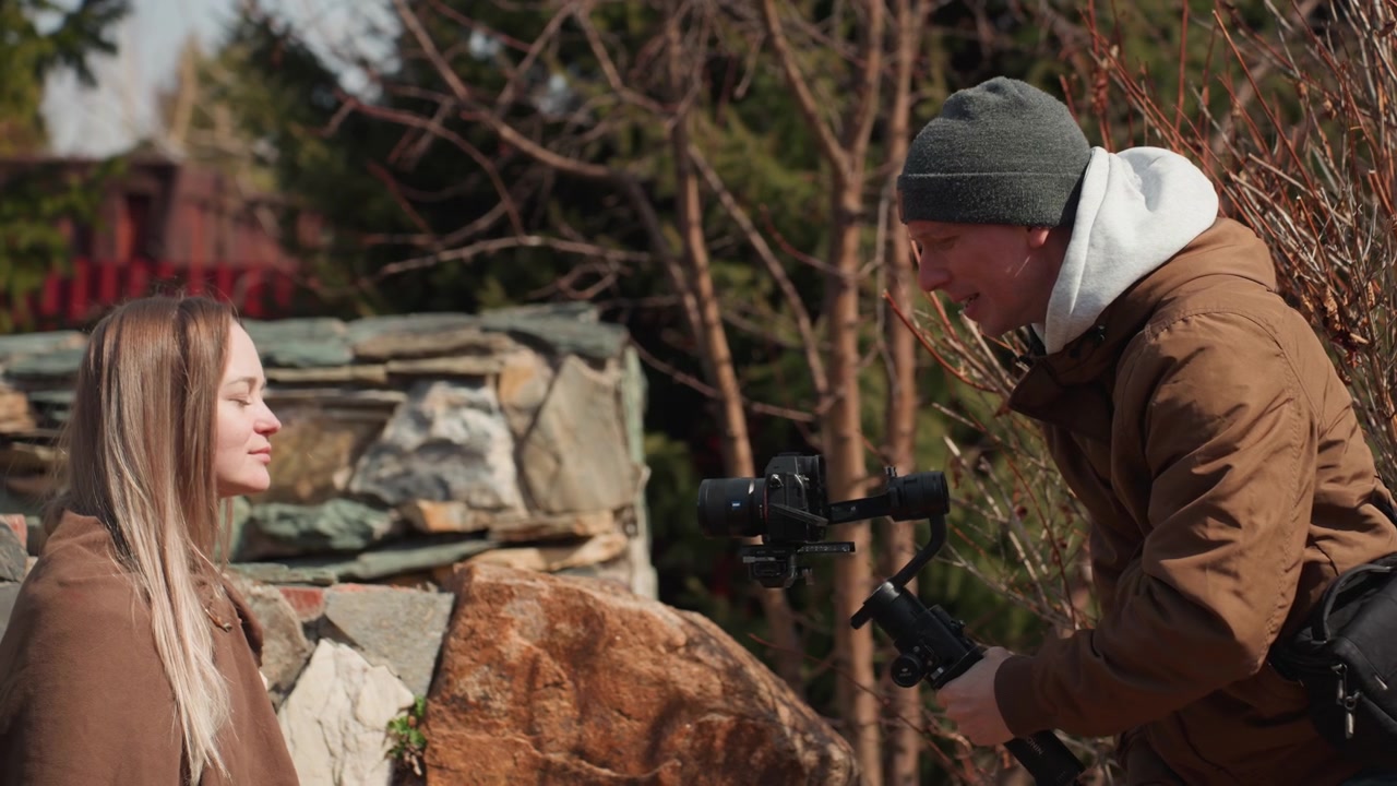 Mand læner sig frem og stabiliserer gimbal for at holde siddende kvinde i syne, mens hun drejer hovedet let og har et roligt, afslappet udtryk, hvilket viser blid bevægelse, kompositionsændring og udendørs produktion