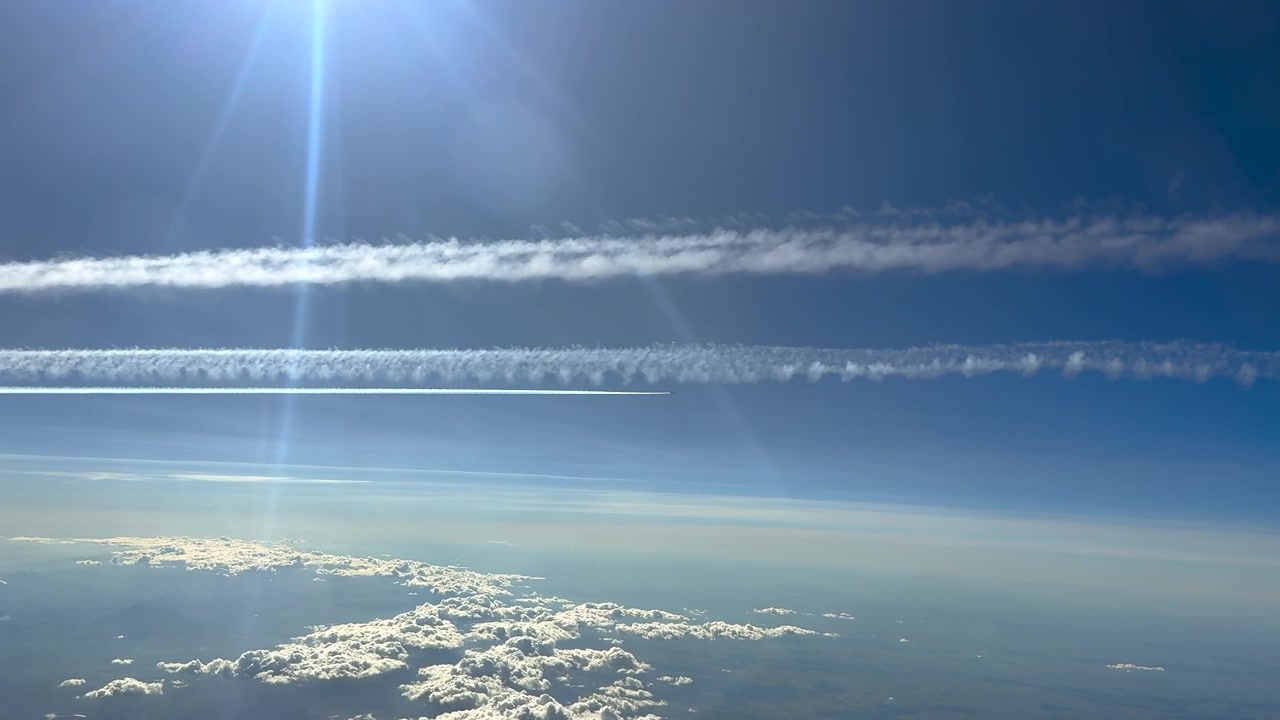 高高度を飛行するジェット機のコックピットから撮影された空撮。青い空に多くの飛行機雲と、光芒を放つ輝く太陽が見える。