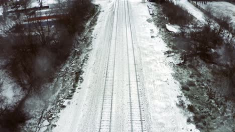 perspective shot of train tracks sandwiched between bare trees and brush and snow flurries blow about