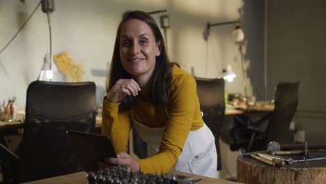 caucasian female jeweller in workshop wearing apron, using tablet, smiling at camera