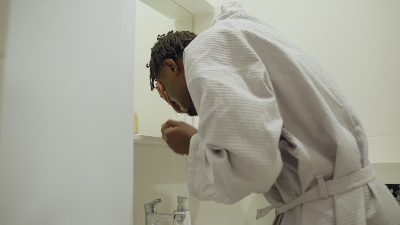 Black youth in bathrobe standing at bathroom sink, bending over, focused, washing hands with soap, modern clean bathroom with soft lighting, morning hygiene routine, self-care, fresh start