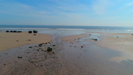 Tiefflugaufnahme-über-Sandstrand-Mit-Felsen-In-Richtung-Des-Ruhigen-Meeres-Mit-Kleinen-Wellen