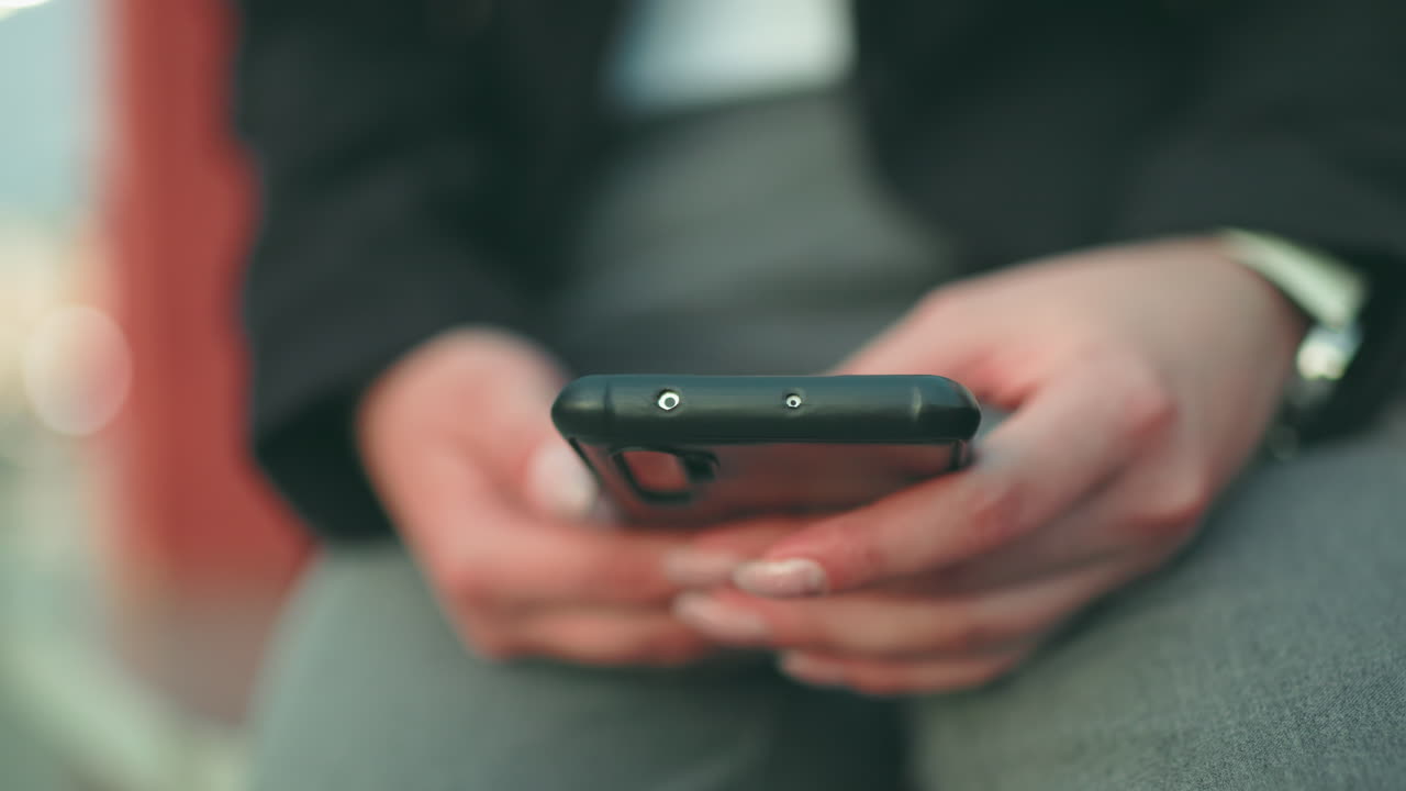 Close up soft focus of young lady with artificial nails operating phone in both hands while seated, wearing black jacket and wristwatch, with soft bokeh lights and blurred background