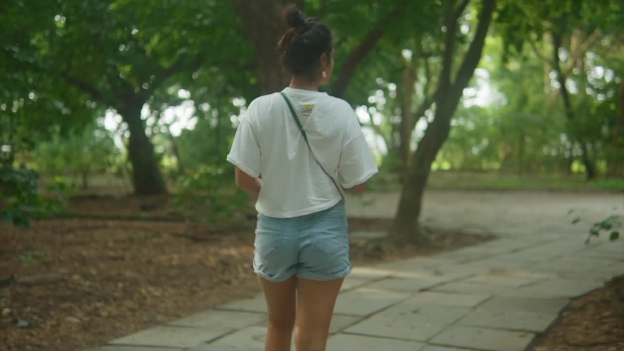 현대적인 옷을 입은 젊은 인도 십대 소녀는 공원에서 푸른 푸른색으로 둘러싸인 보행로에서 자연을 즐기고 있습니다.