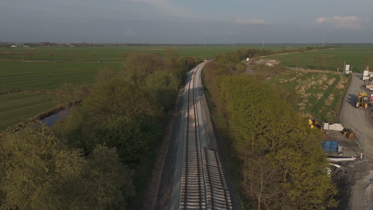 Aerial shot of a railway construction site in rural East Frisia, near the new Friesenbrücke project. Tracks being laid with heavy machinery and materials. Spring landscape in evening light. Shot in 4K
