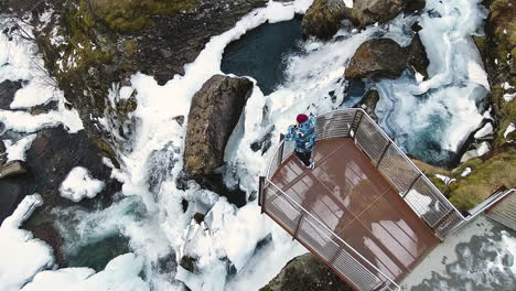 man on observation platform in steep cliff with overview of icy waterfall in town of geiranger in norway during winter season