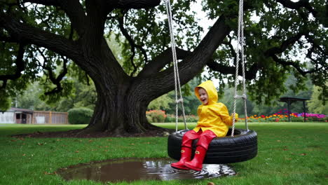 a child in a yellow raincoat and red boots playing on a tire swing over a puddle