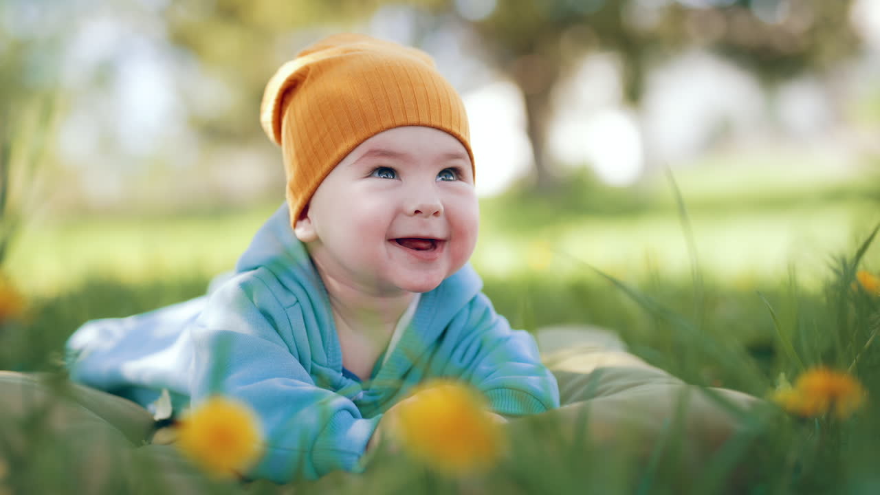 Baby boy in orange hat lies in nature. Kid looks up with surprise, smiles sweetly and then starts to yawn.