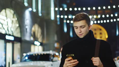 A-young-man-walks-through-the-night-city-and-uses-a-smartphone