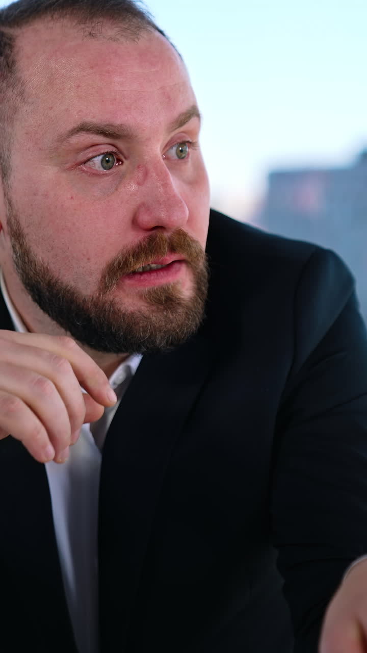 Portrait of a tired businessman at workplace. Bearded office worker in suit sitting at the table with a laptop on window background in the city. Vertical video