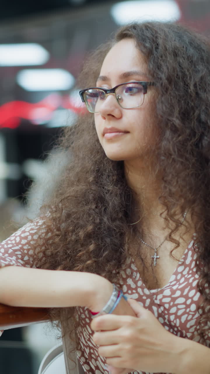 Young adult in a patterned dress gazes thoughtfully into the distance with one hand resting on a railing, background features people browsing and shopping, with vibrant neon signs and sale posters