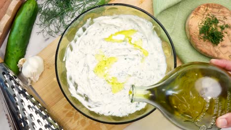 making tzatziki greek or mediterranean sauce or dip for a party. dip or sauce in a green bowl on a wooden cutting board with dill, green napkin and a grater. adding olive oil to the tzatziki sauce.