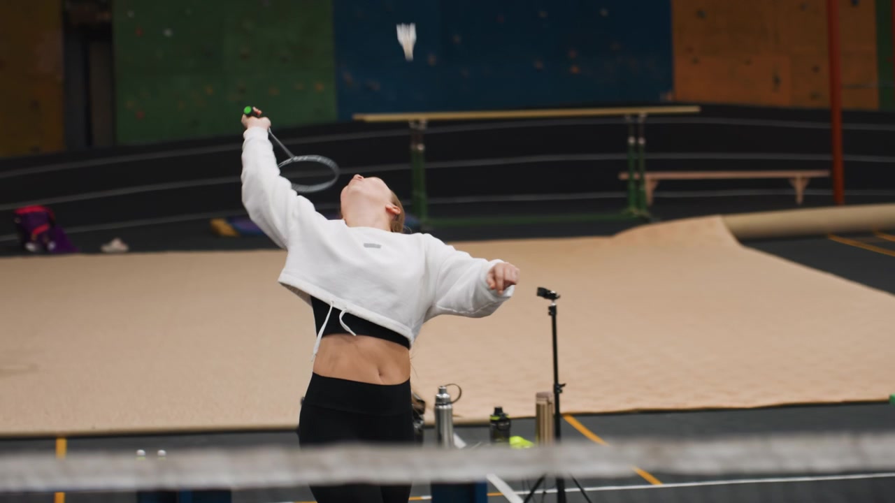 Smiling white woman playing badminton on indoor court near net, recreational player warming up with racket and shuttlecock, relaxed sweatshirt, light drills, laughter and upbeat energy, emphasis