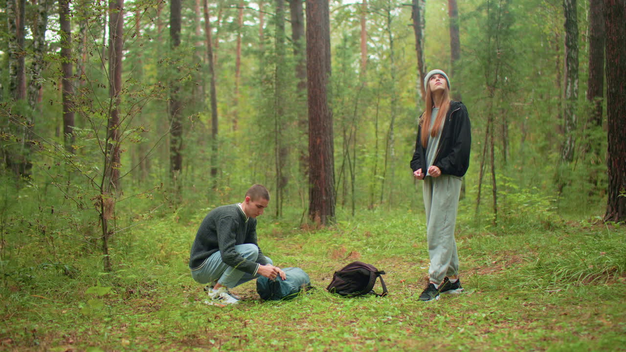 traveler in forest as man kneels to unzip backpack lying on ground while woman in casual outfit stands nearby looking around thoughtfully, surrounded by pine trees and lush greenery