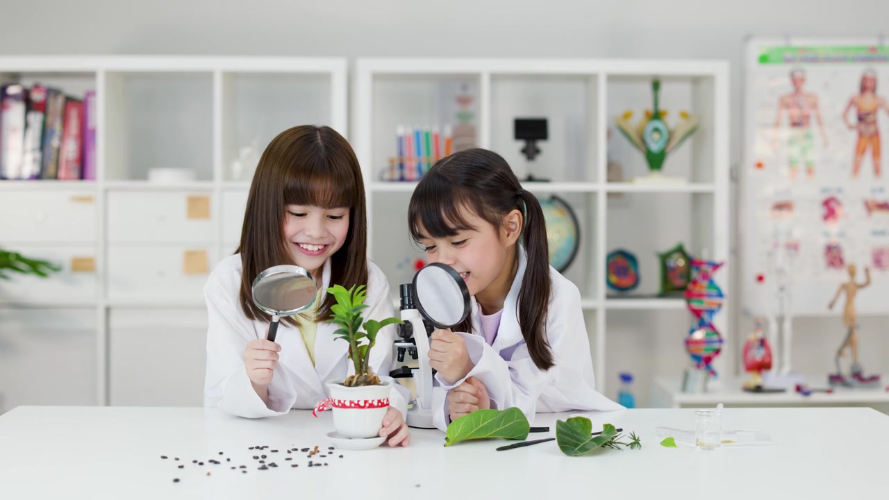 Two young girls examine potted plants and seeds using magnifying glasses in a bright classroom.
