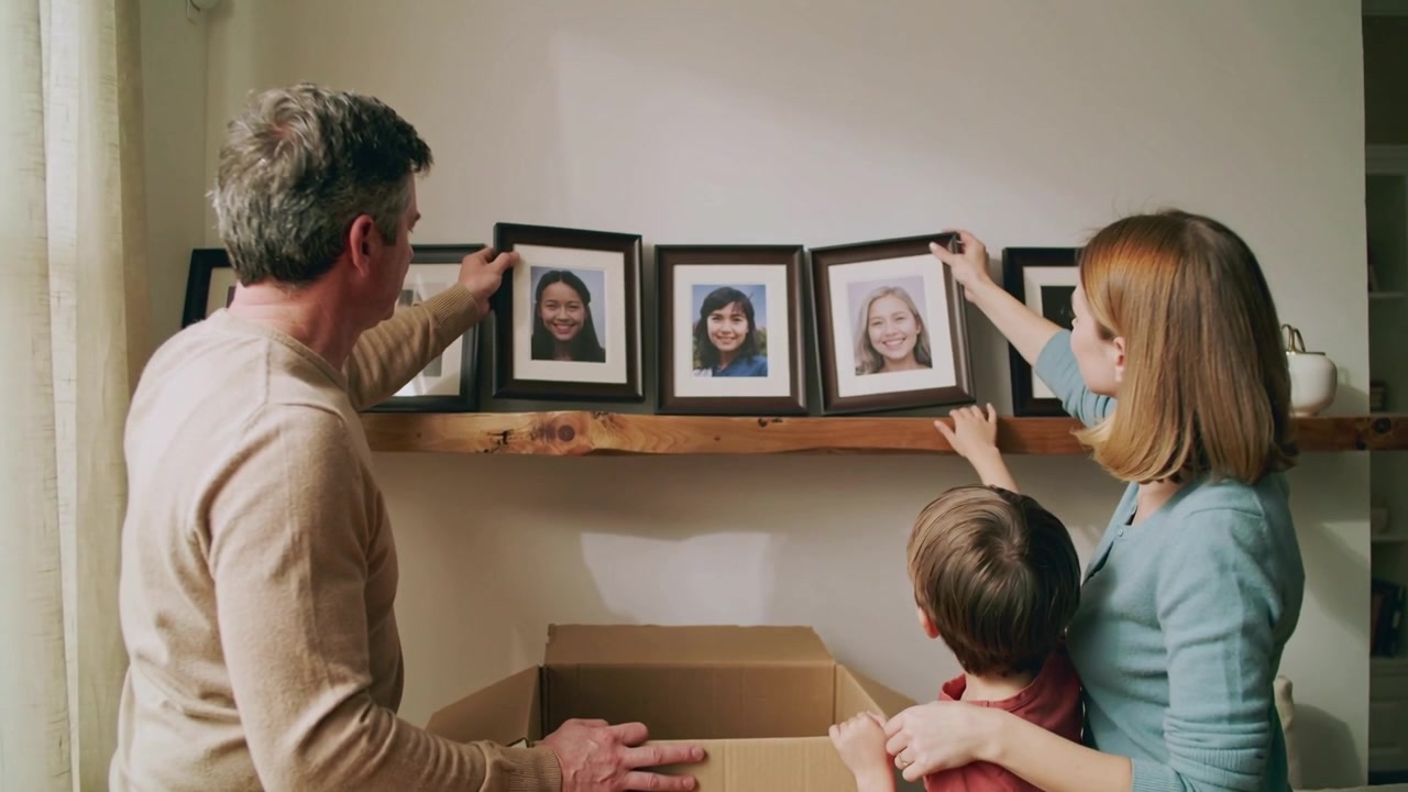 Unpacking parents in beige and blue placing frames on wooden shelf with open box, son watching