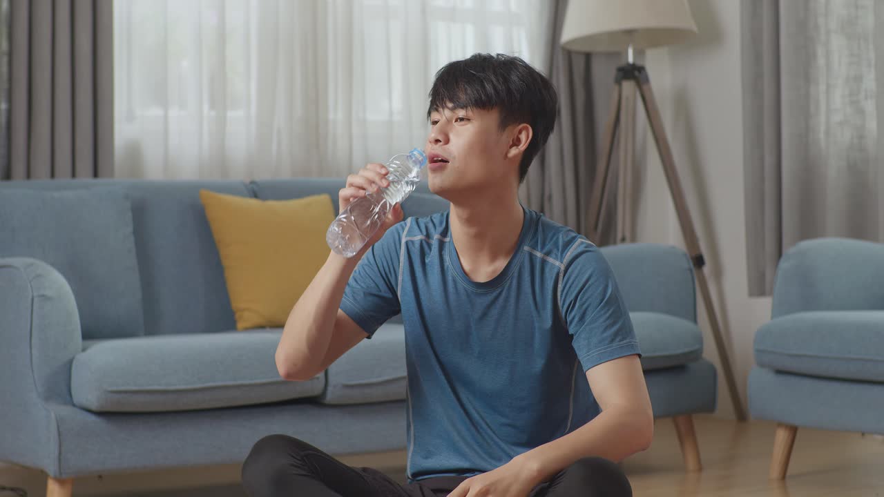 스포츠 옷을 입은 운동체조의 아시아인 남자가 아침 운동을 마친 후 집에서 미니멀한 인테리어를 가진 넓고 밝은 아파트에서 물을 마시고 있습니다.