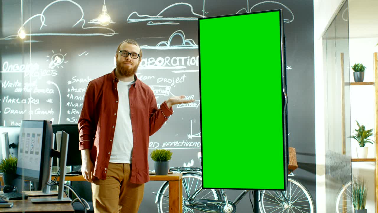 남성 개발자는 모의 녹색 화면으로 인터랙티브 화이트 보드를 보며, 프레젠테이션 제스처와 미소로 화면을 가리키며, 배경에는 프로젝트 계획이 그려진 칠판 벽이 있는 크리에이티브 오피스입니다.