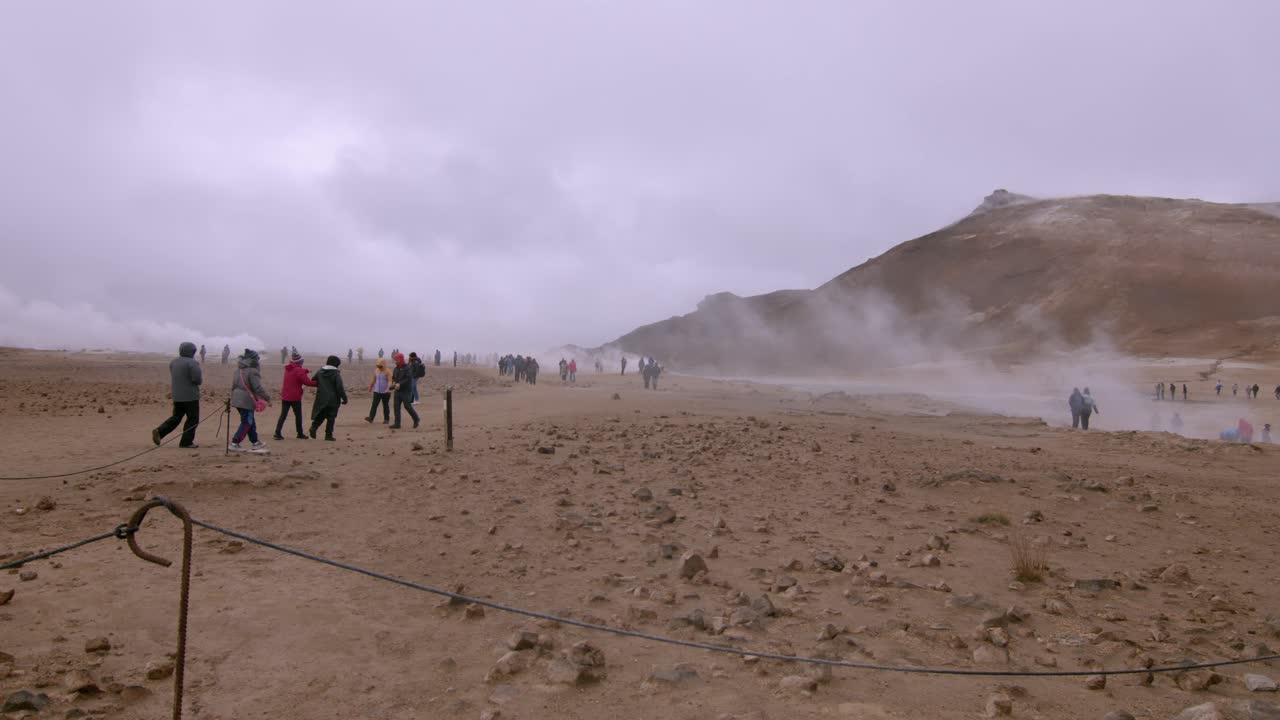 Tourists explore a steamy geothermal field under a cloudy sky with mountains in the background