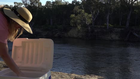 blonde woman checking what's in the esky cooler box on river edge camping